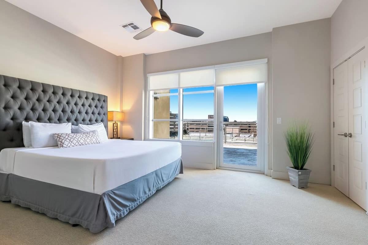 A bedroom with a large bed, a ceiling fan, a view of the ocean through a sliding glass door, and a potted plant.