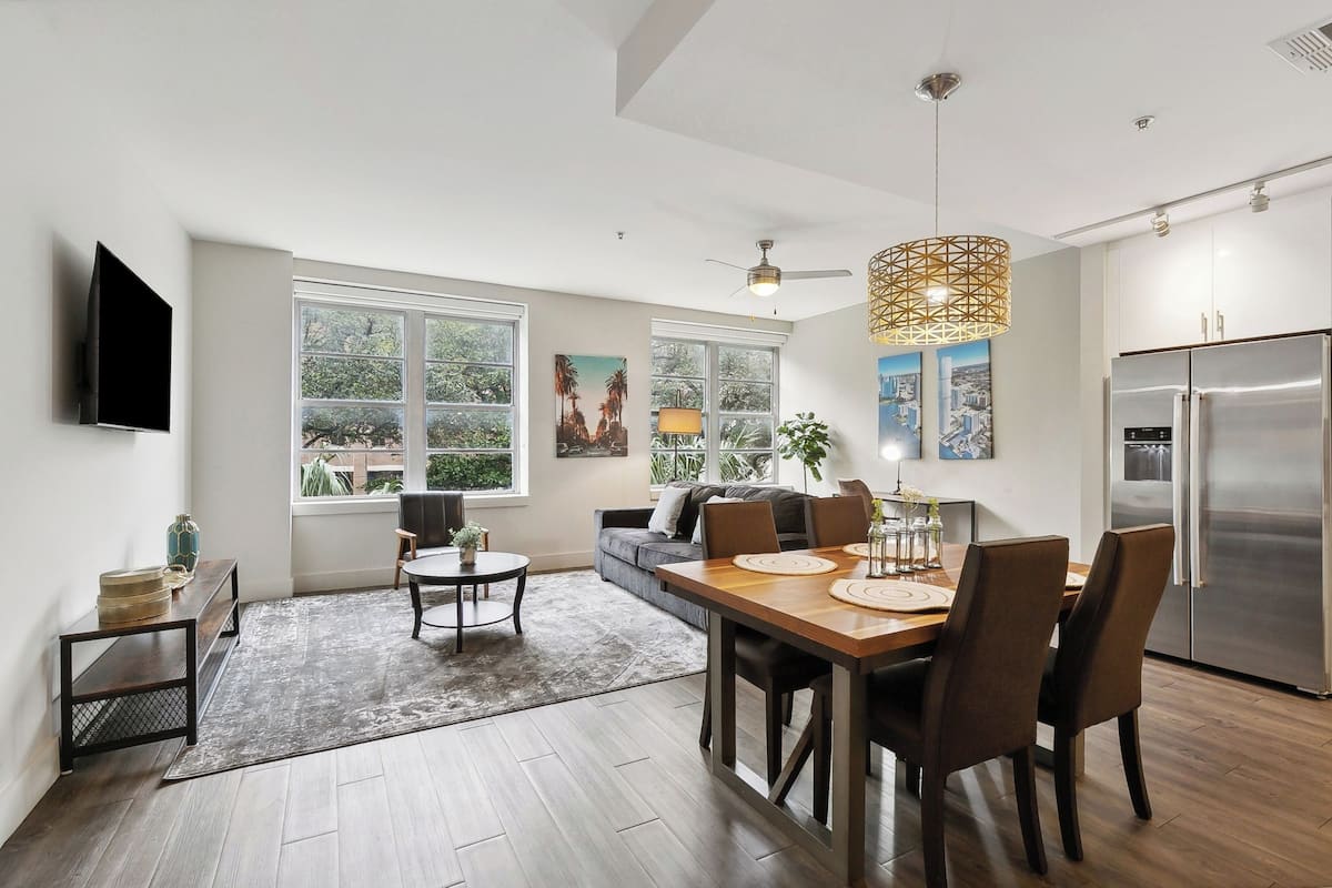 A modern kitchen with a built-in refrigerator, a dining area with a wooden table and chairs, and large windows offering a view of greenery outside.