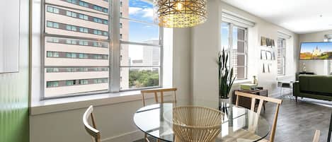 Un comedor moderno con una mesa de vidrio y sillas de mimbre, ventanas amplias con vistas a la ciudad, y una lámpara colgante.