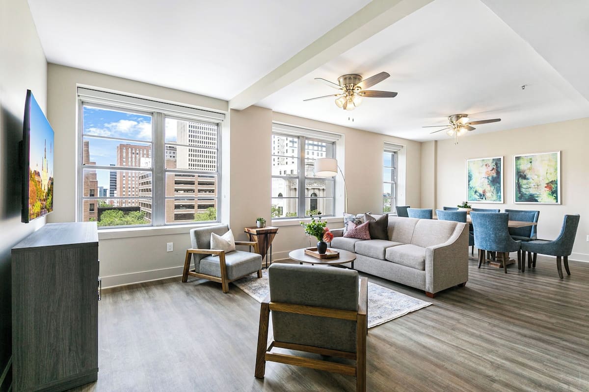 A spacious living room with a large window, a ceiling fan, a sofa, a coffee table, and a dining area with blue chairs.