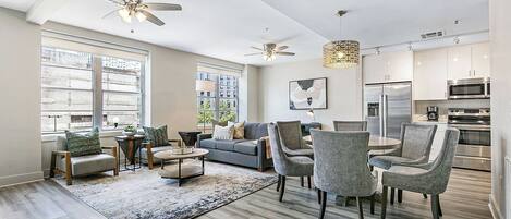 Una sala de estar moderna con un sofá gris, sillones y una mesa de centro. Hay ventiladores de techo, una araña y un ventanal con vistas a edificios.