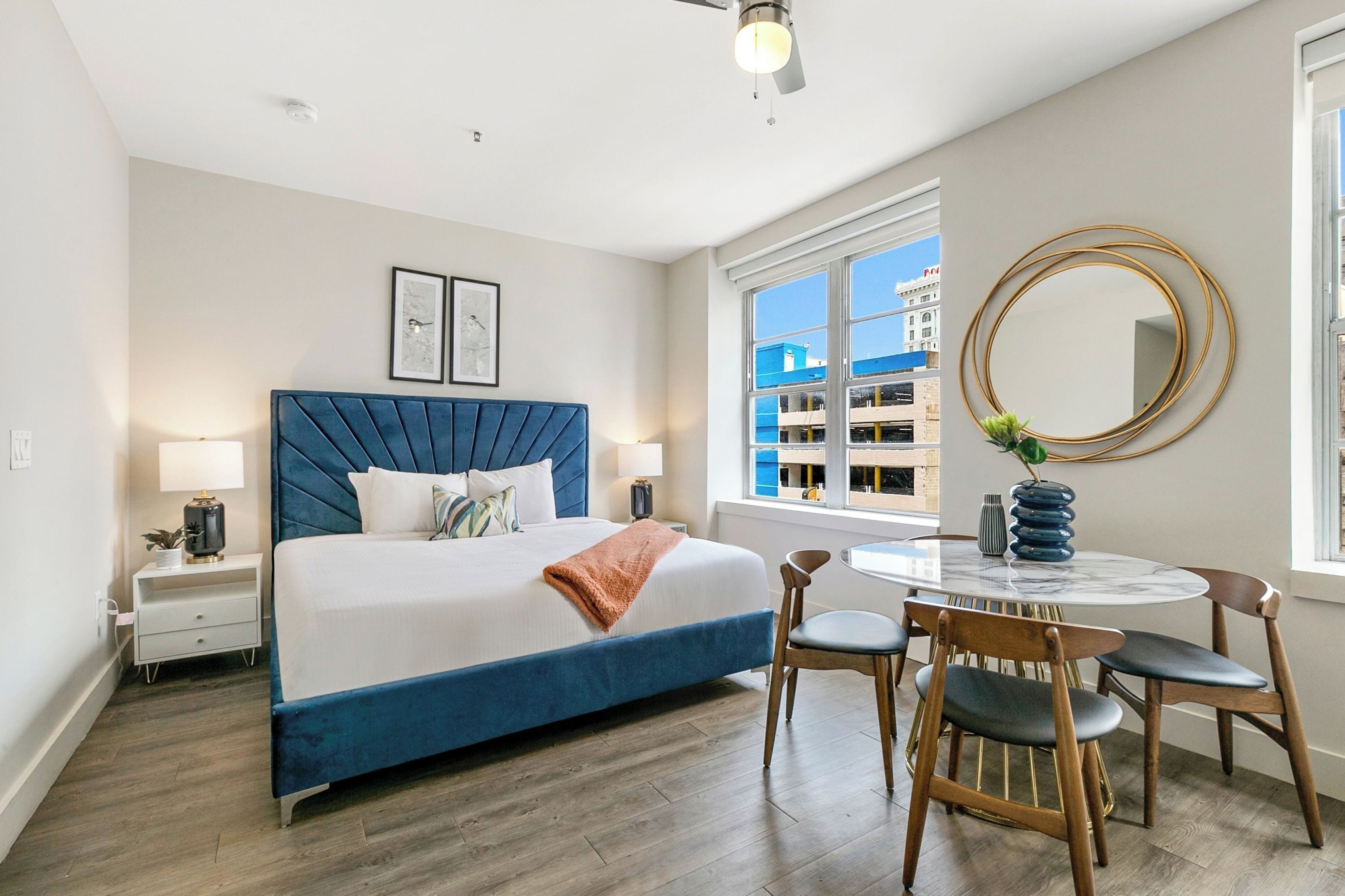 A modern bedroom with a large bed, bedside tables, a round dining table, and a view of buildings outside the window.
