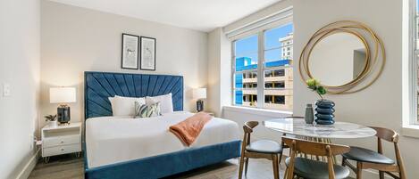 A modern bedroom with a large bed, bedside tables, a round dining table, and a view of buildings outside the window.
