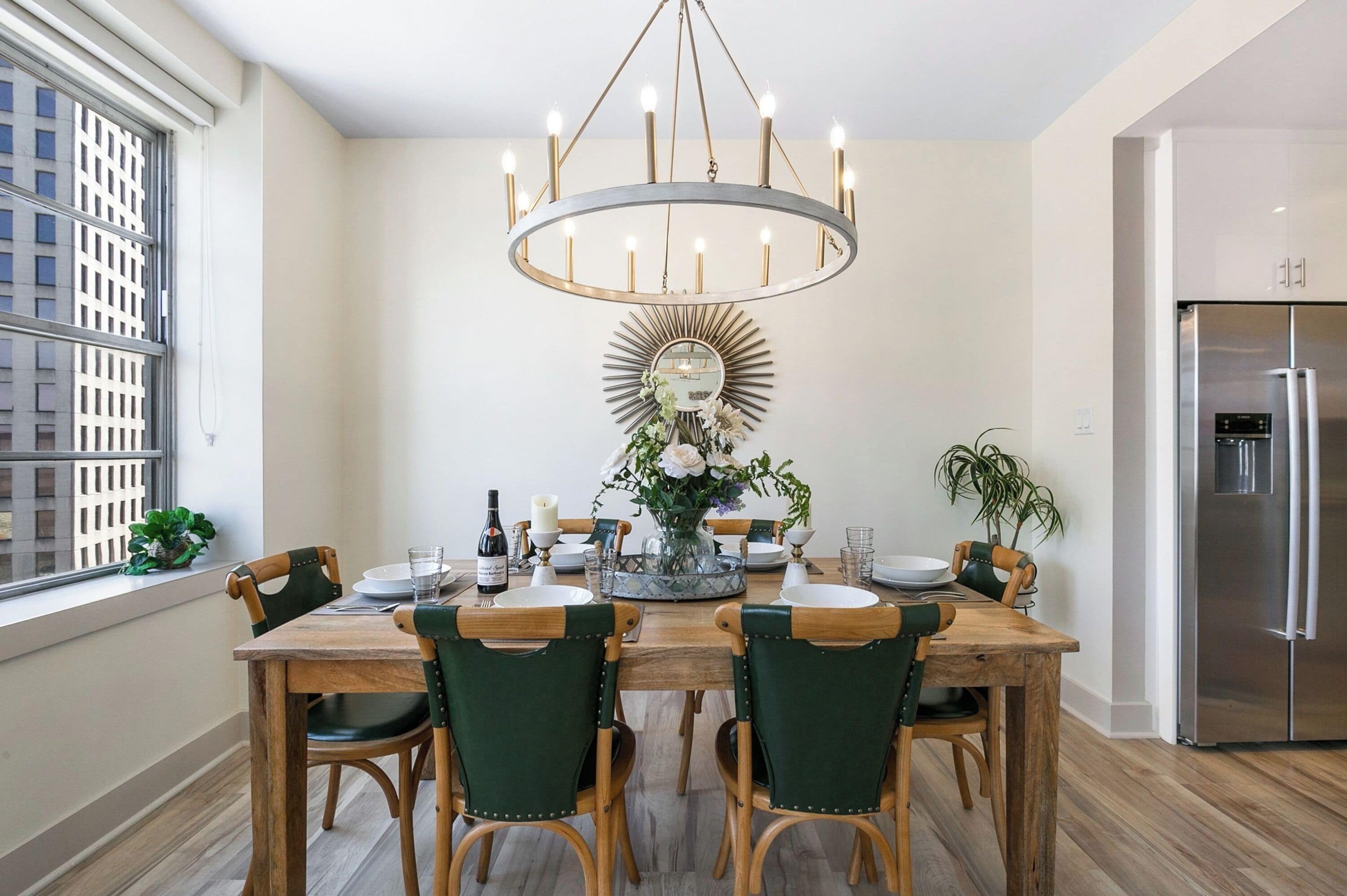 Un comedor con una mesa de madera, sillas tapizadas en verde, una araña, un espejo con marco de sol y un refrigerador de acero inoxidable.