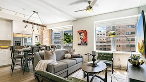 A modern living room with a grey sofa, green armchairs, a dining area with a table and chairs, and a kitchen in the background. - California Building (New Orleans)