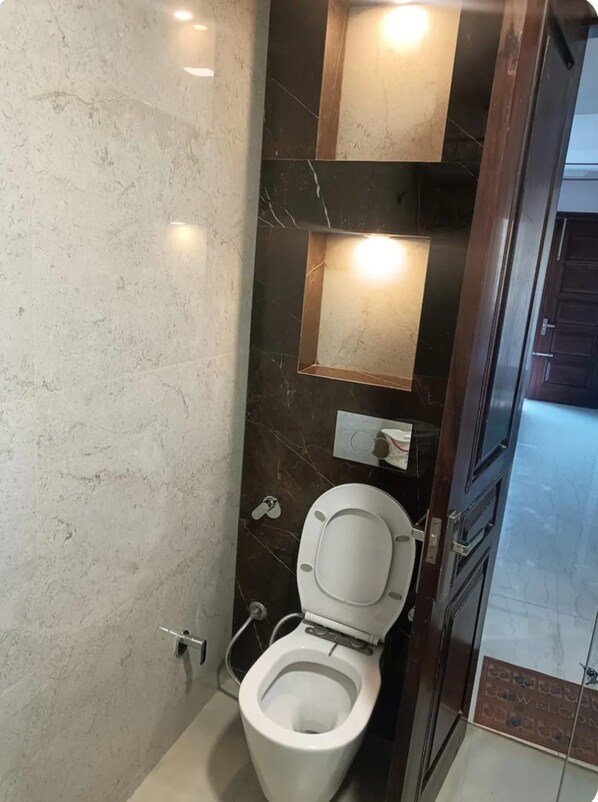 A modern bathroom with a marble wall, a white toilet, and a wall-mounted light fixture. - HomiStay - Luxury Room (Panchkula)