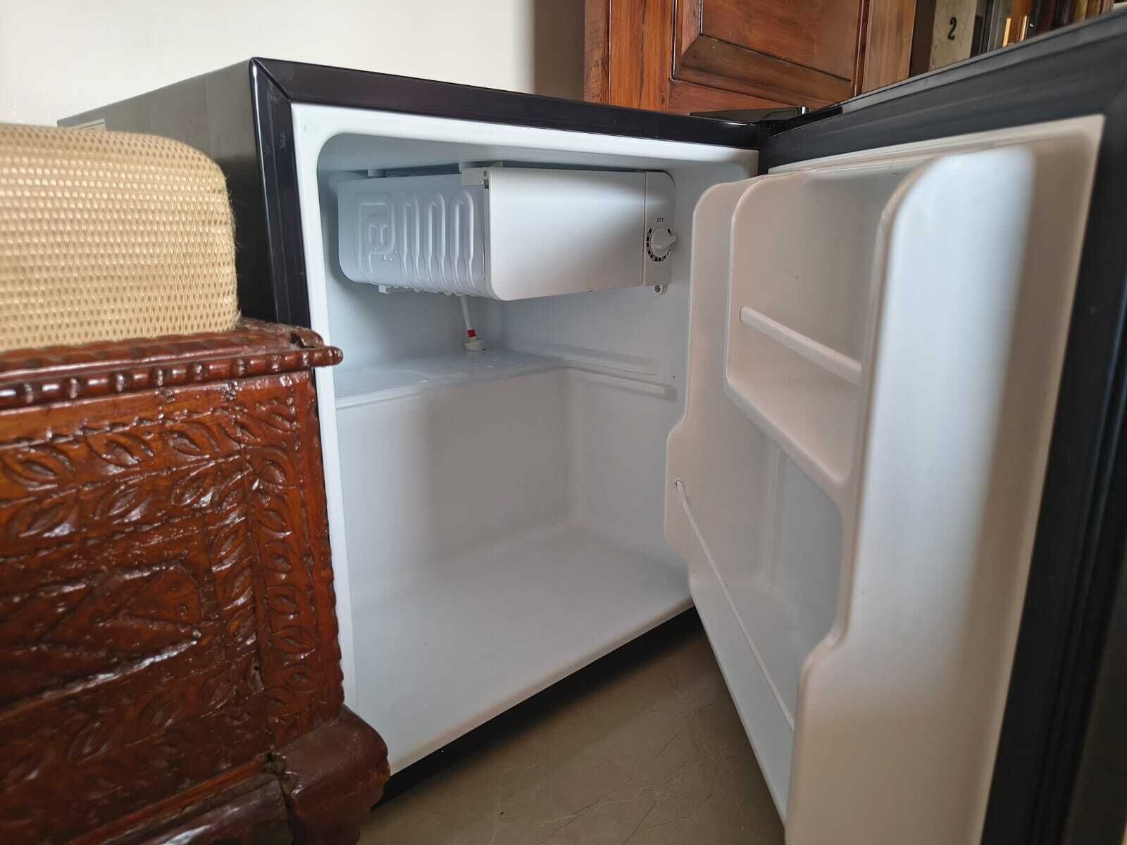 A compact refrigerator with a freezer compartment, situated in a kitchen with wooden cabinetry and a textured beige rug.