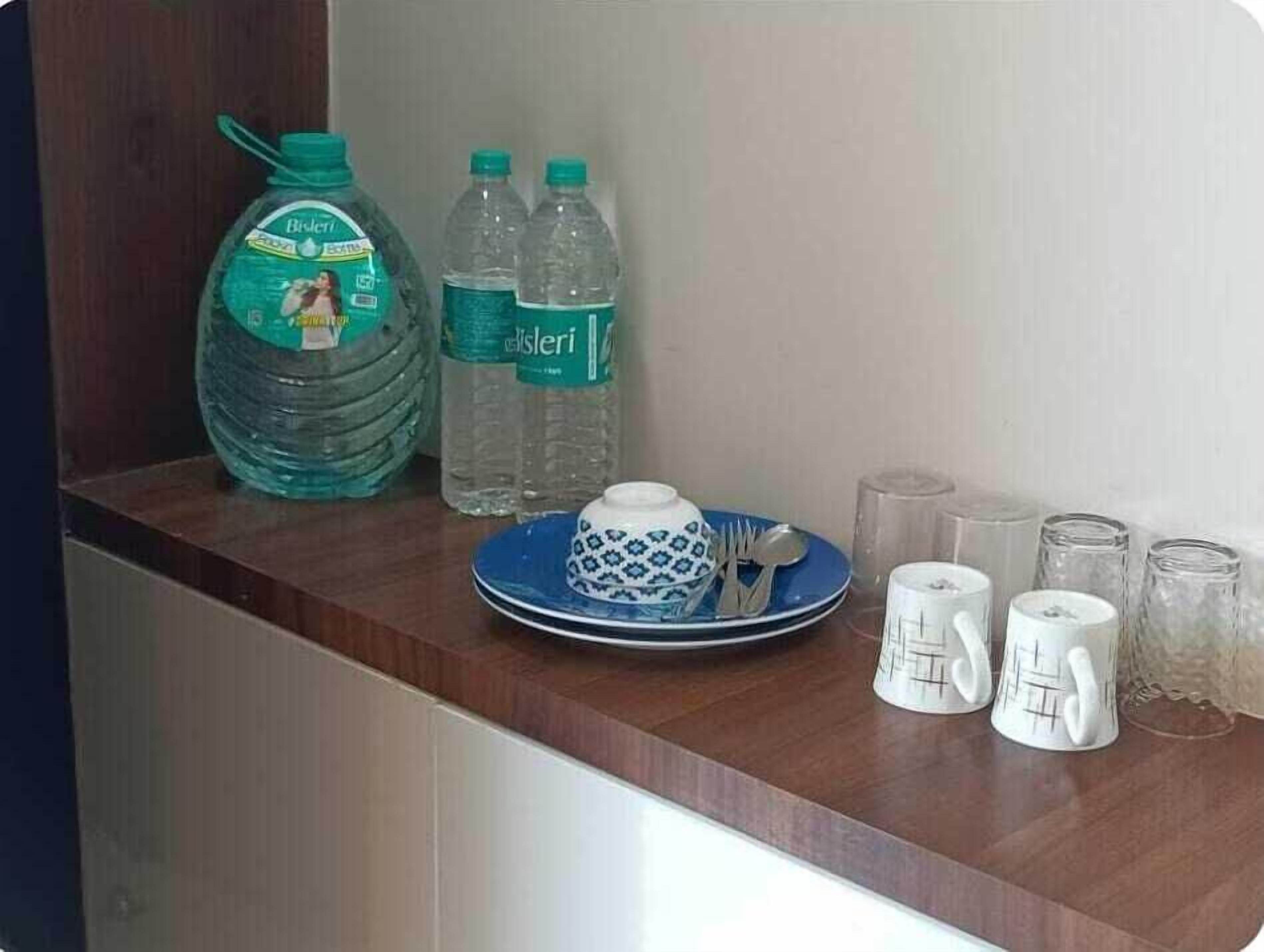 Una encimera de cocina con un dispensador de agua grande, dos botellas de agua, un plato azul con un cuenco blanco, una cuchara, un tenedor y varios frascos de vidrio.