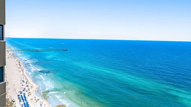Una playa con aguas azules cristalinas, un muelle que se adentra en el mar y un edificio con balcones con vistas a la playa.