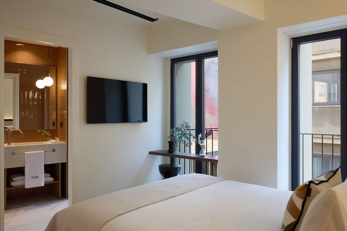 A modern hotel room with a large bed, a flat-screen TV, a bathroom with a mirror and sink, and a balcony with a plant.
