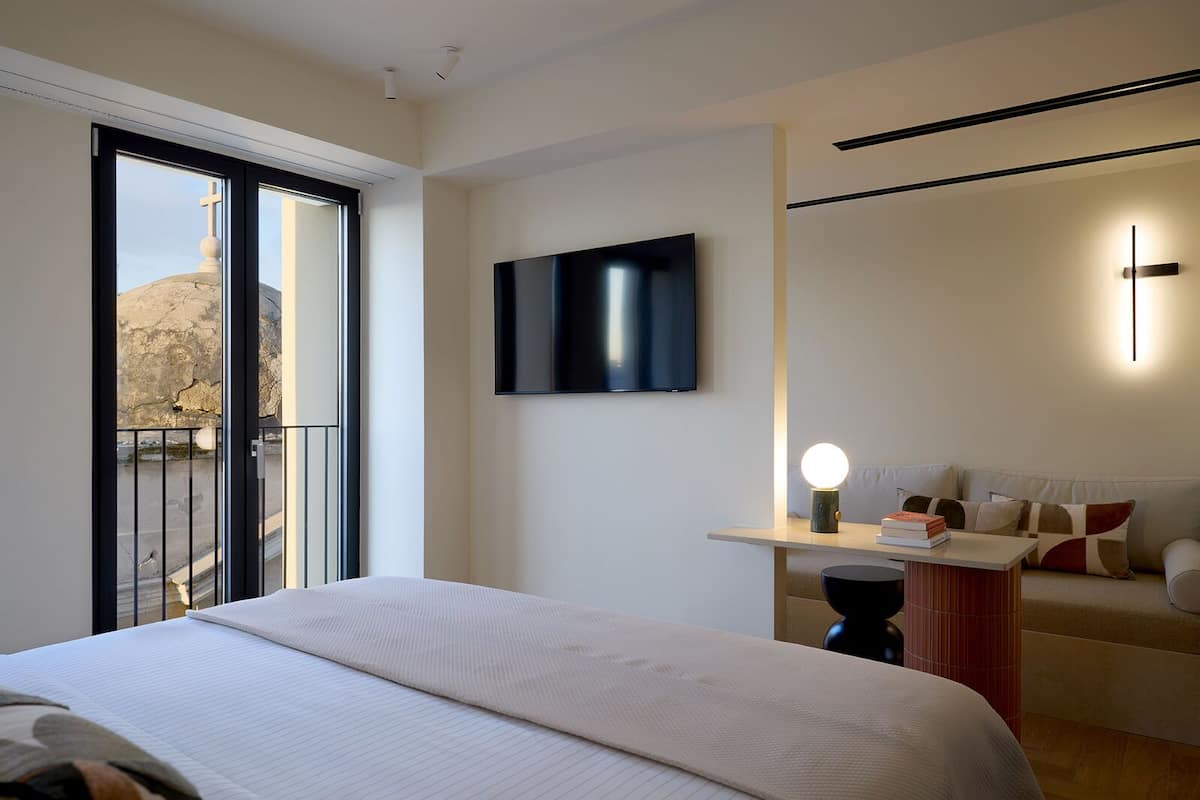 A modern hotel room with a large bed, a flat-screen TV, a bedside table with a lamp, and a view of the outdoors through a sliding door.