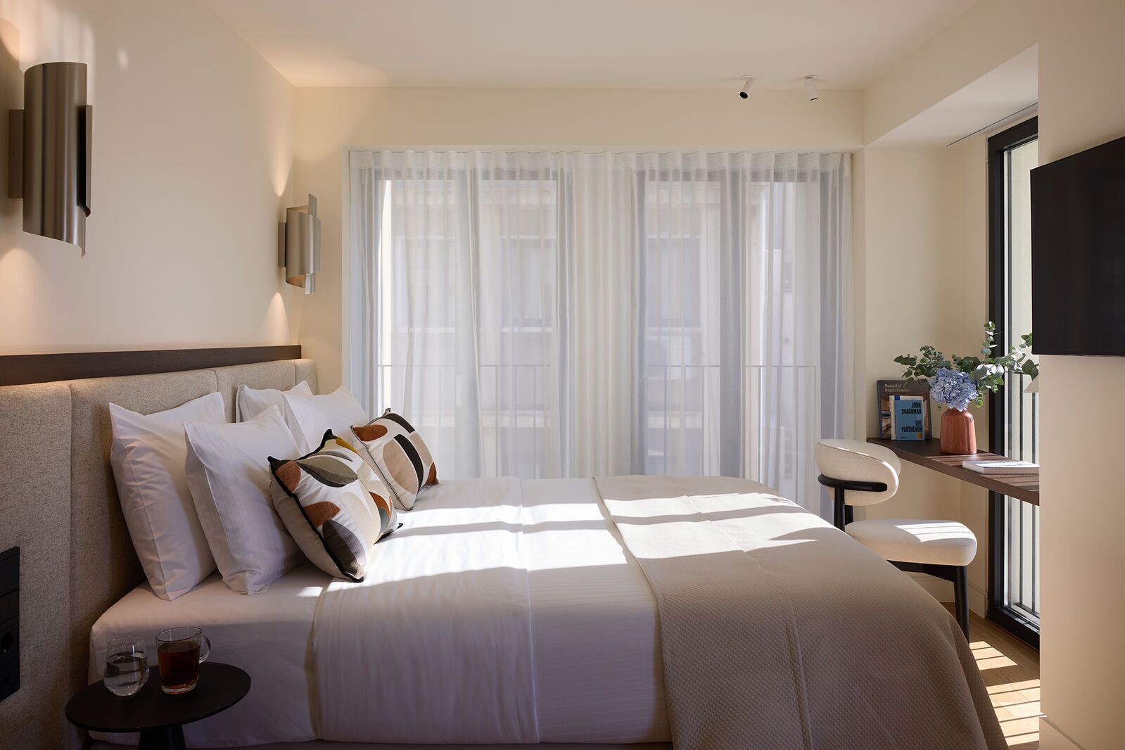 Ein modernes Hotelzimmer mit einem großen Bett, einem Nachttisch, einem Sessel, einem Fenster mit durchsichtigen Vorhängen und einem an der Wand befestigten Fernseher.