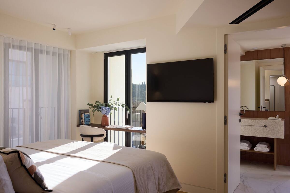 A modern hotel room with a large bed, a desk with a chair, a TV mounted on the wall, and a bathroom with a mirror and towel rack.