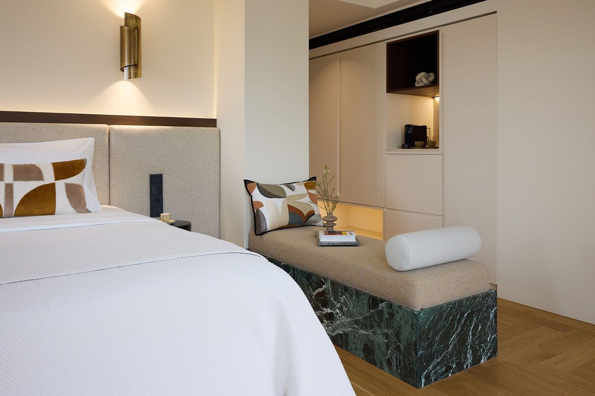 A modern hotel room with a large bed, a nightstand, a built-in bench with a vase, and a wall-mounted shelf with decorative items.