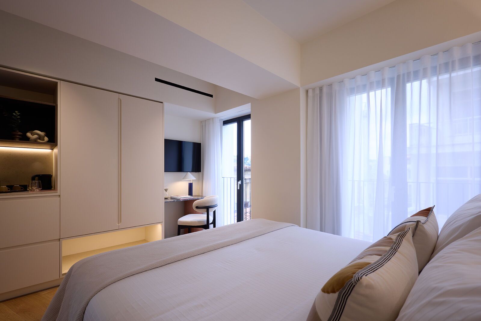 A modern hotel room with a large bed, a desk with a chair, and a view through sheer curtains.