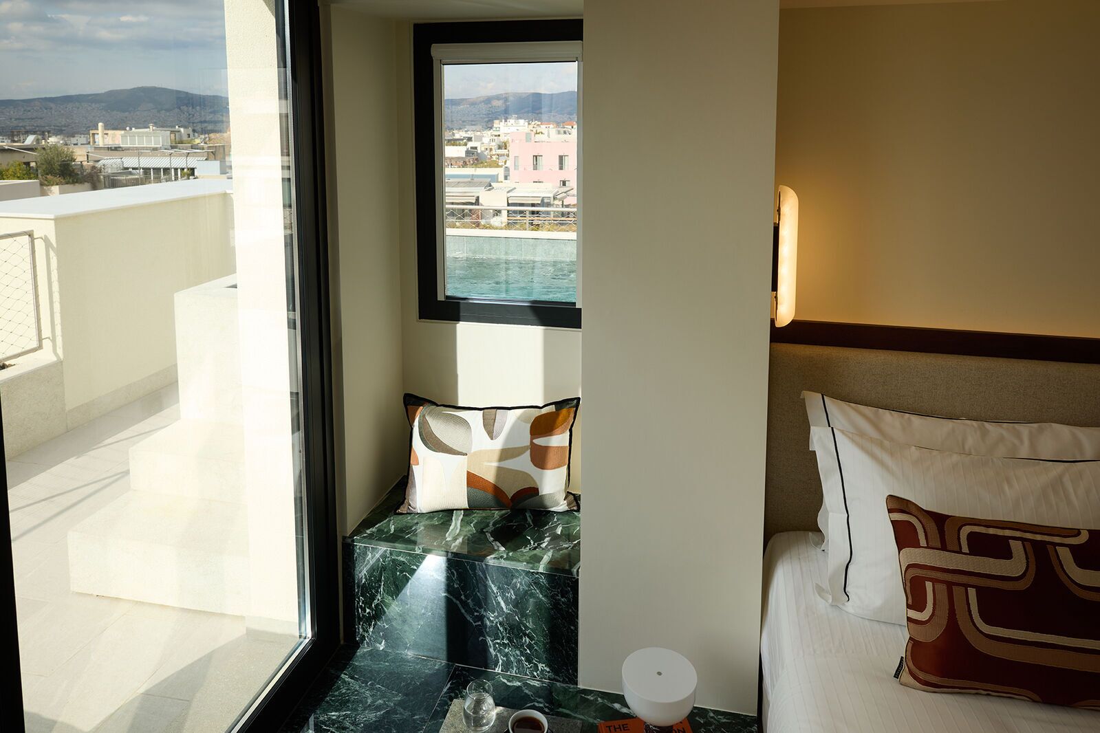 Une chambre d’hôtel avec un balcon, une vue sur la ville, une table recouverte de carreaux de marbre et un lit agrémenté d’oreillers à motifs.