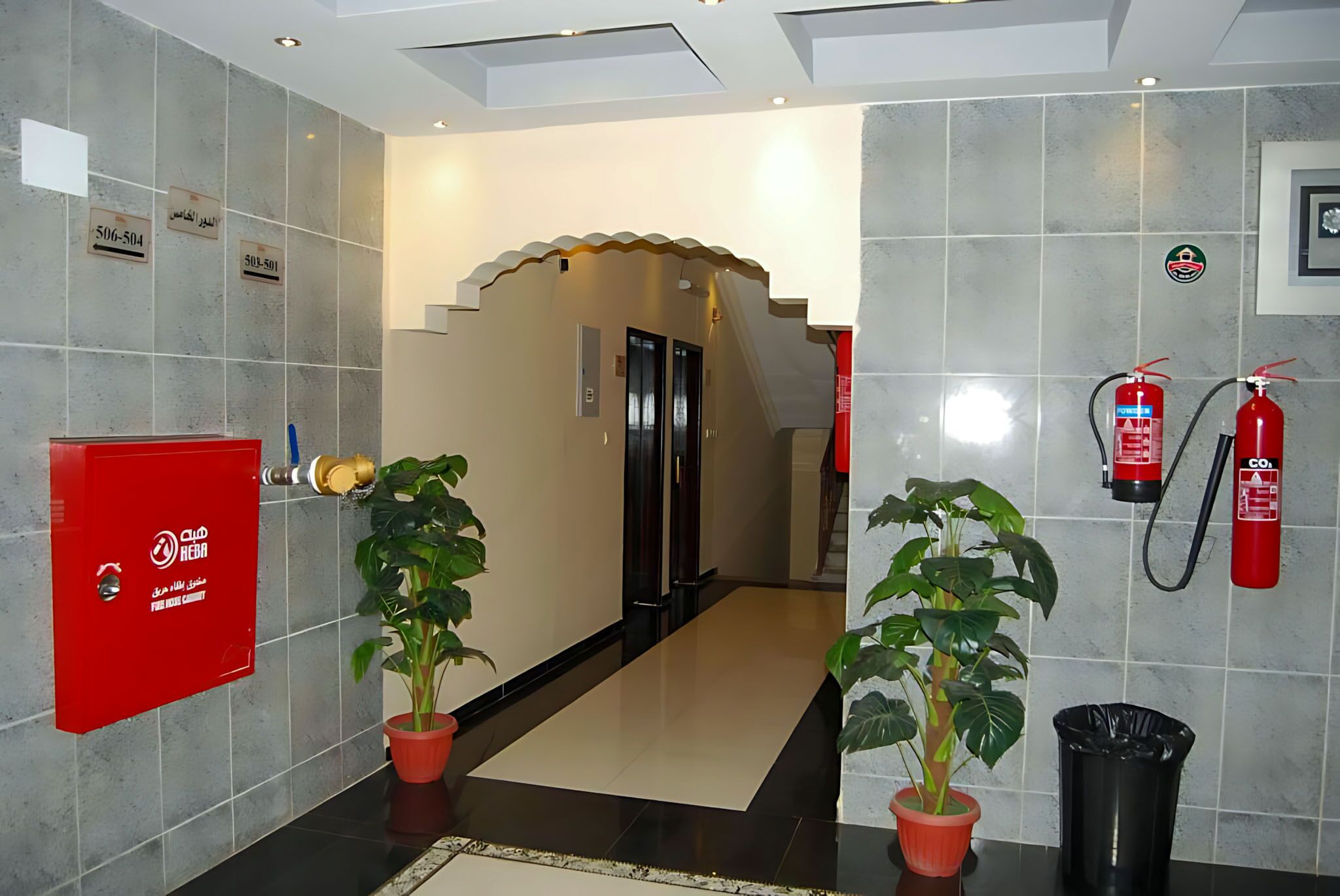 A hotel lobby with a tiled floor, a reception desk, potted plants, a fire extinguisher, and a staircase leading to the rooms.