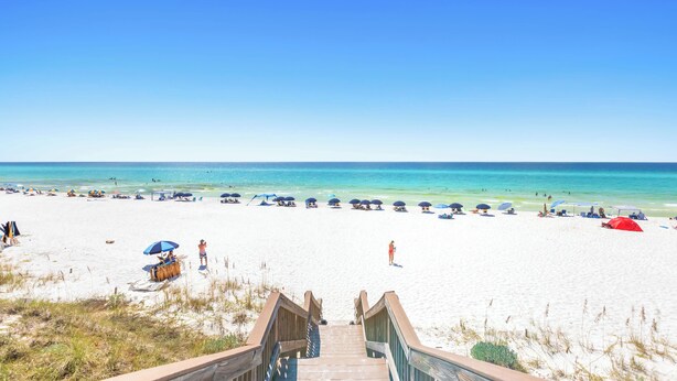Ein Strand mit einer Holztreppe, die zum Sand führt, Sonnenschirmen und Personen.