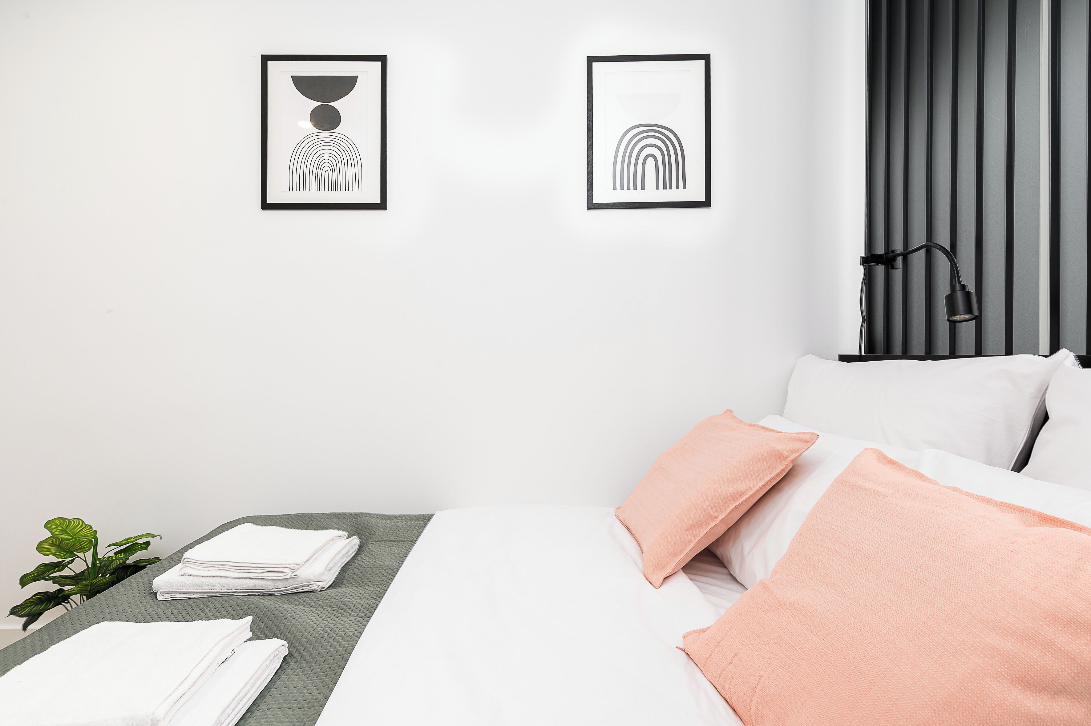 Una habitación de hotel con cama, almohadas, una mesita de noche con una planta y dos cuadros en la pared.
