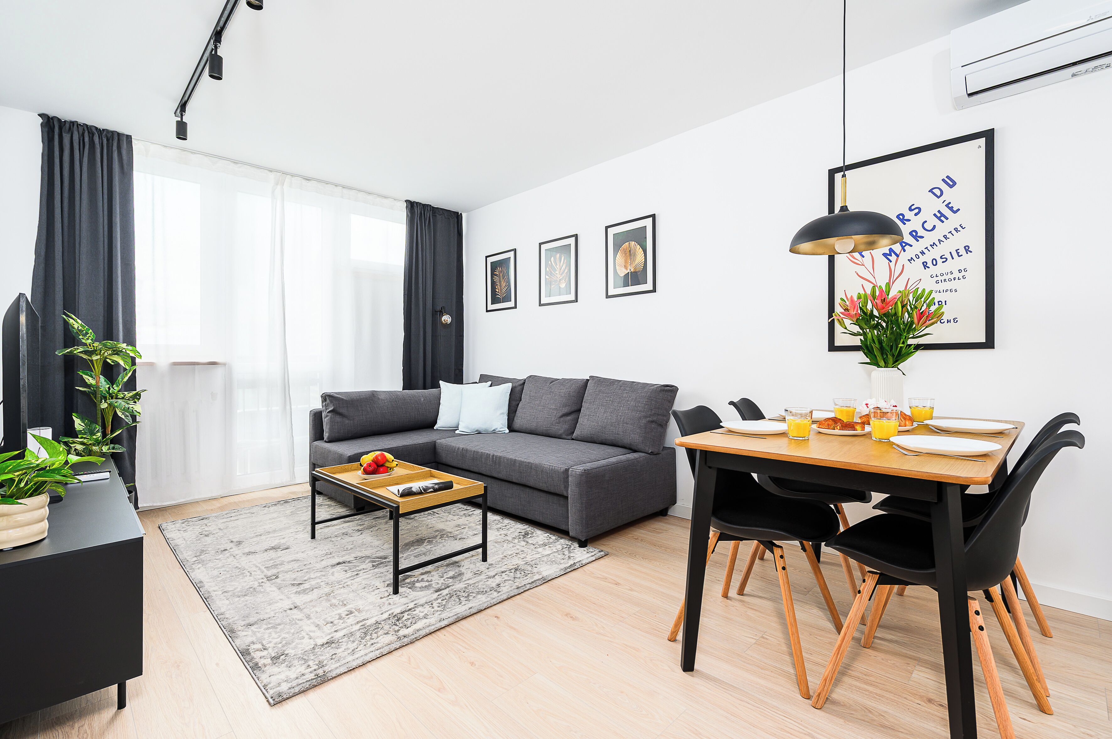 A modern living room with a grey sofa, a dining table with chairs, and a coffee table. There are framed pictures on the wall and a potted plant on the table.