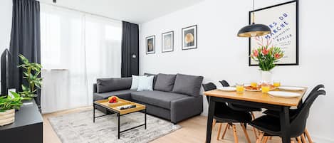 A modern living room with a grey sofa, a dining table with chairs, and a coffee table. There are framed pictures on the wall and a potted plant on the table.