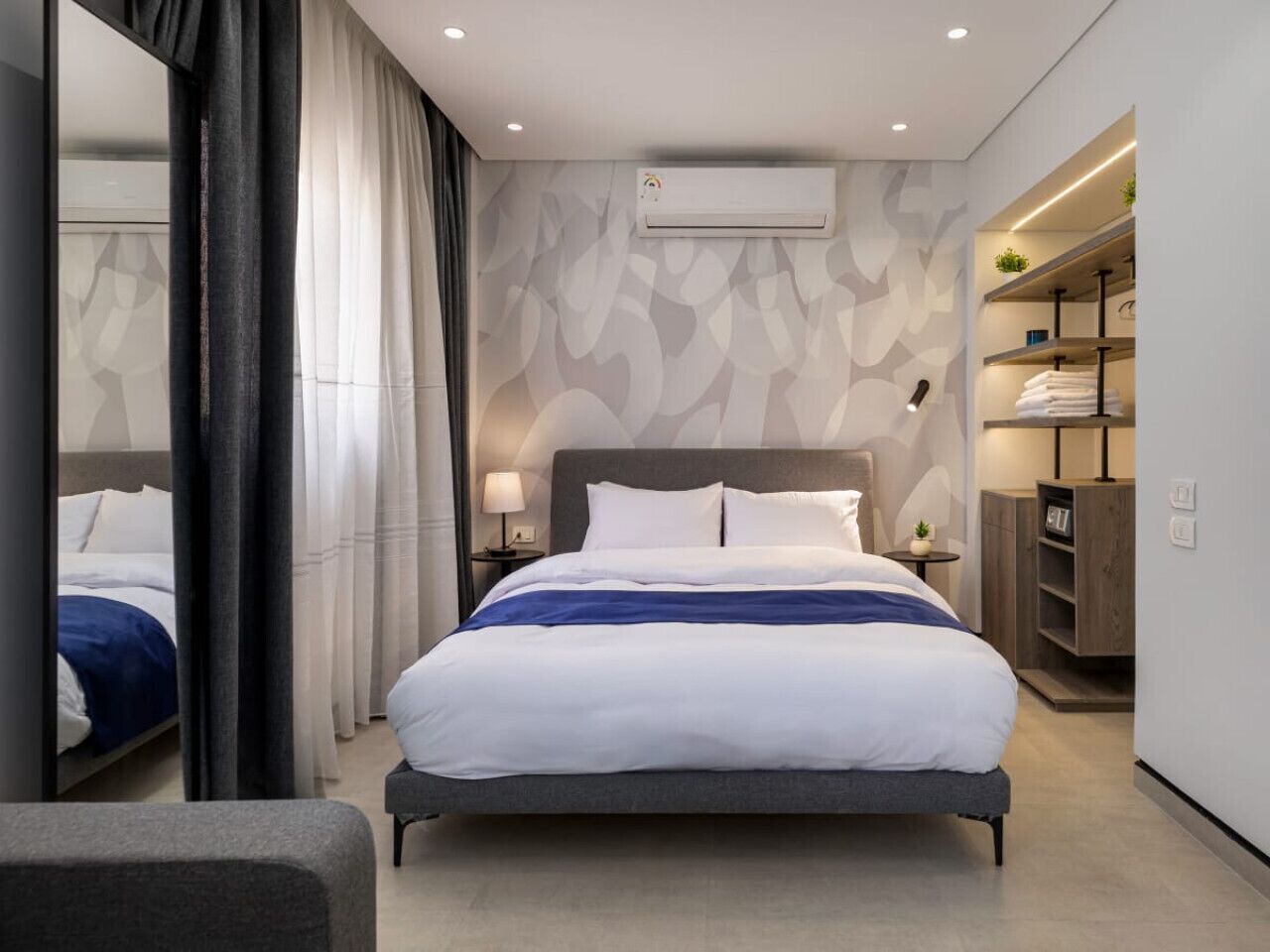 Moderni hotellihuone, jossa on suuri sänky, yöpöydät, ilmastointilaite ja sisäänrakennettu vaatekaappi.