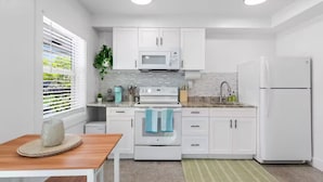 Una cocina moderna con armarios blancos, microondas, estufa y refrigerador. Hay una ventana con persianas, una planta y una pequeña mesa con un tapete.