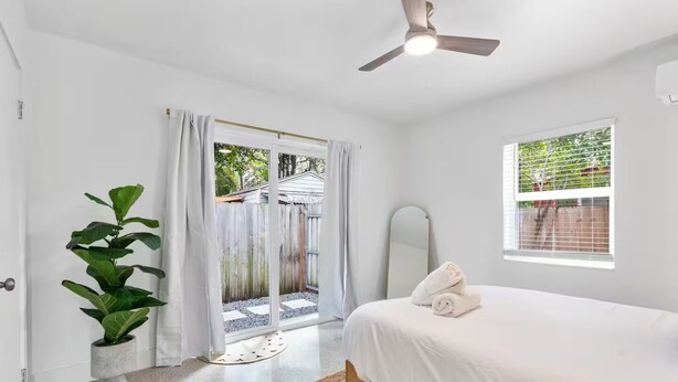 Un dormitorio con cama, ventilador de techo, ventana y puerta corredera que da a un espacio al aire libre.