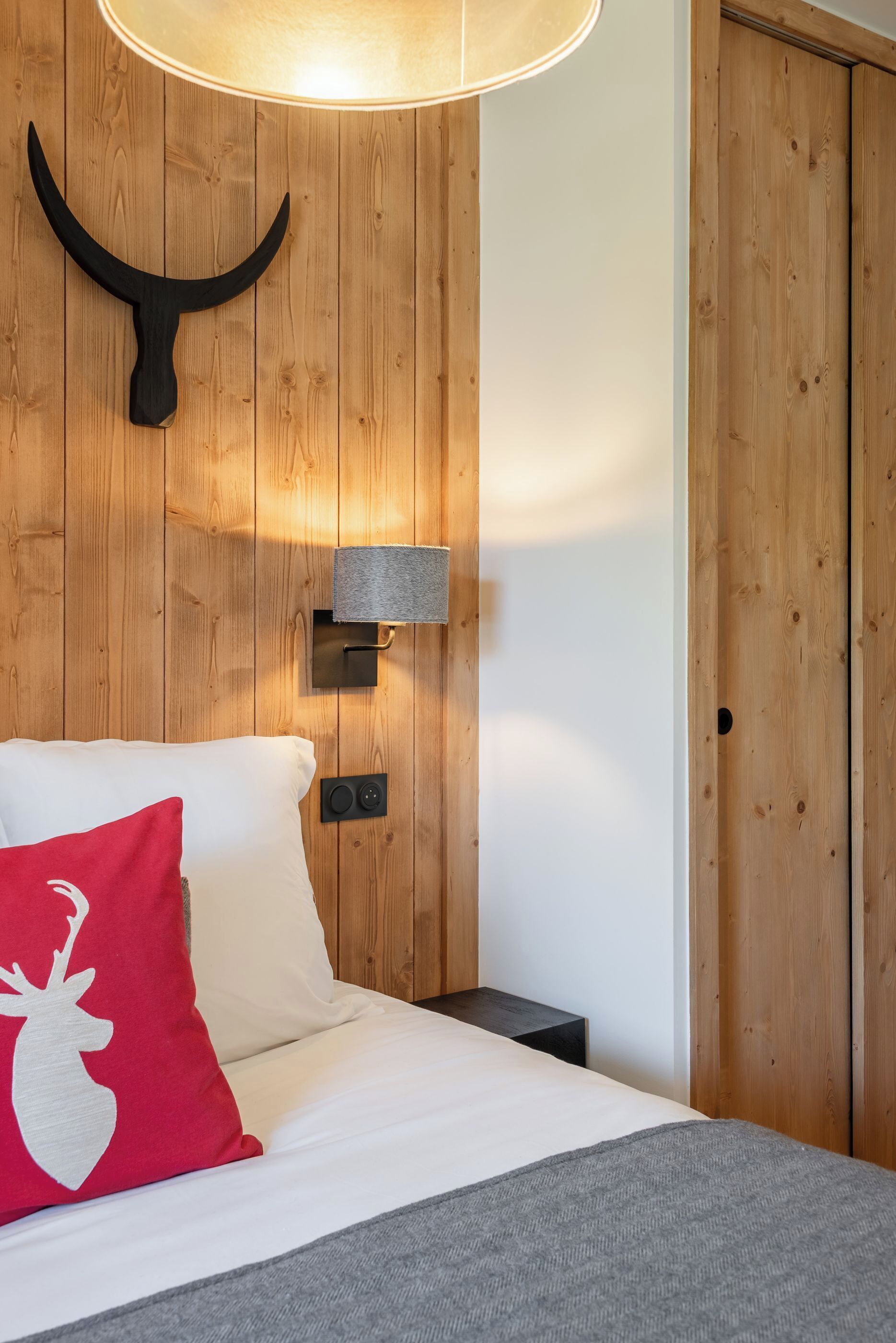 A bedroom with a wooden wall, a bed with white and red pillows, a mounted horn decoration, a wall-mounted lamp, and a wooden wardrobe.