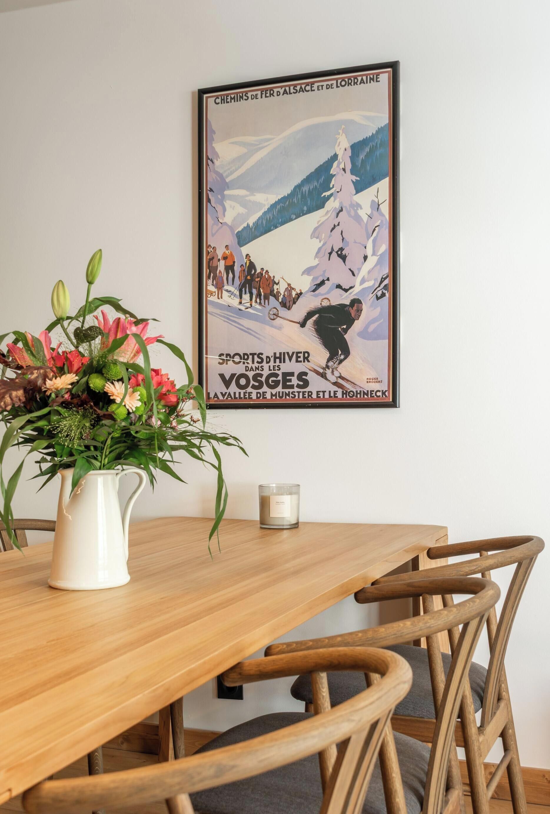 A dining table with a white pitcher of flowers, a candle, and wooden chairs.