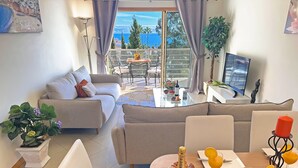 Een moderne woonkamer met een bank, een eettafel met eten en drinken, en een balkon met uitzicht op zee.