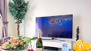 Een kamer met een televisie waarop een mooi uitzicht te zien is, een fles Moët & Chandon champagne, verse aardbeien, bosbessen, een bloemenvaas en een bord met gebak en jus d'orange.