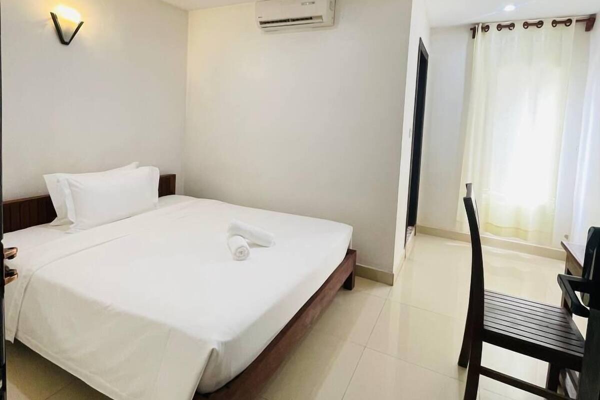 A hotel room with a bed, a chair, an air conditioning unit, and a wall-mounted light fixture.