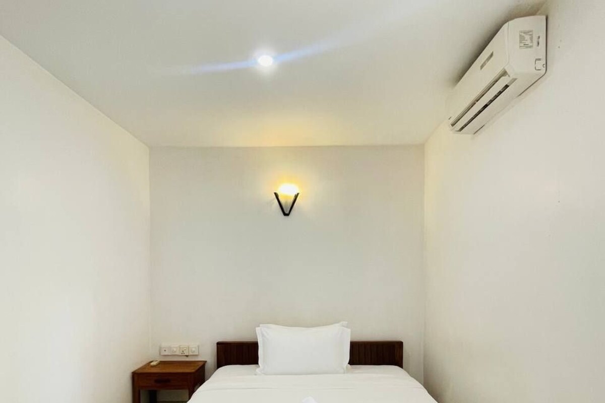 A neatly made bed with white linens, a bedside table with a lamp, and a wall-mounted air conditioner.