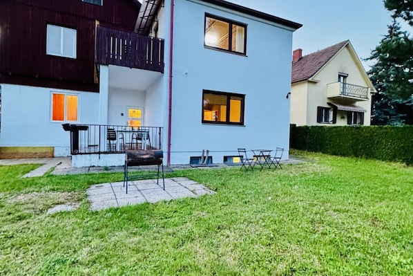 Ein zweistöckiges Haus mit einem Balkon, einer Terrasse mit Stühlen und einem gepflegten Rasen.