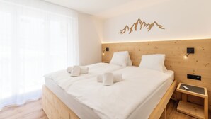 A modern hotel room with a wooden headboard, a large bed with white linens, a bedside table with a remote control, and a wall-mounted light fixture. - Alpin Apartment bei der Bergbahn (Zermatt)