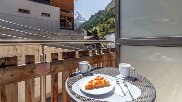 A dining table with a slice of pizza and two white mugs, overlooking a mountainous landscape. - Alpin Apartment bei der Bergbahn (Zermatt)