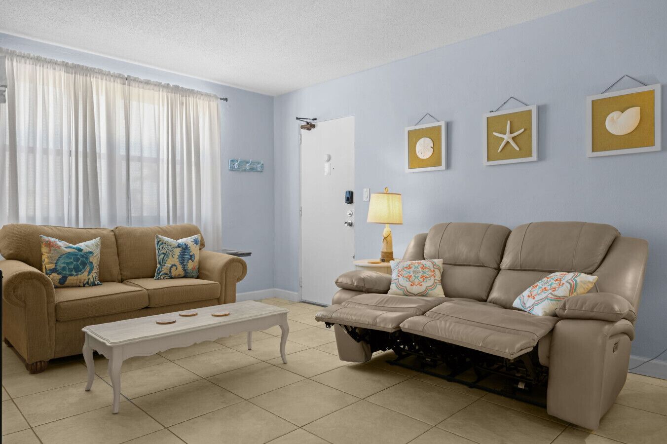 Un salon avec un canapé beige, une table basse blanche et un fauteuil inclinable. Les murs sont décorés, notamment avec une étoile de mer et un cœur.