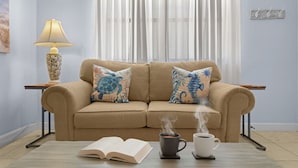 A living room with a beige sofa, two decorative pillows, a table with a book and two cups of coffee, a lamp, and a window with sheer curtains. - Beachside Getaway: 2-Bed Condo with Pool, Walk to St. Pete Sand (St. Pete Beach/Tierra Verde)