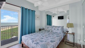 Ein Schlafzimmer mit Bett, Deckenventilator, Nachttisch, Lampe, Fernseher und einem Balkon mit Blick auf den Strand.