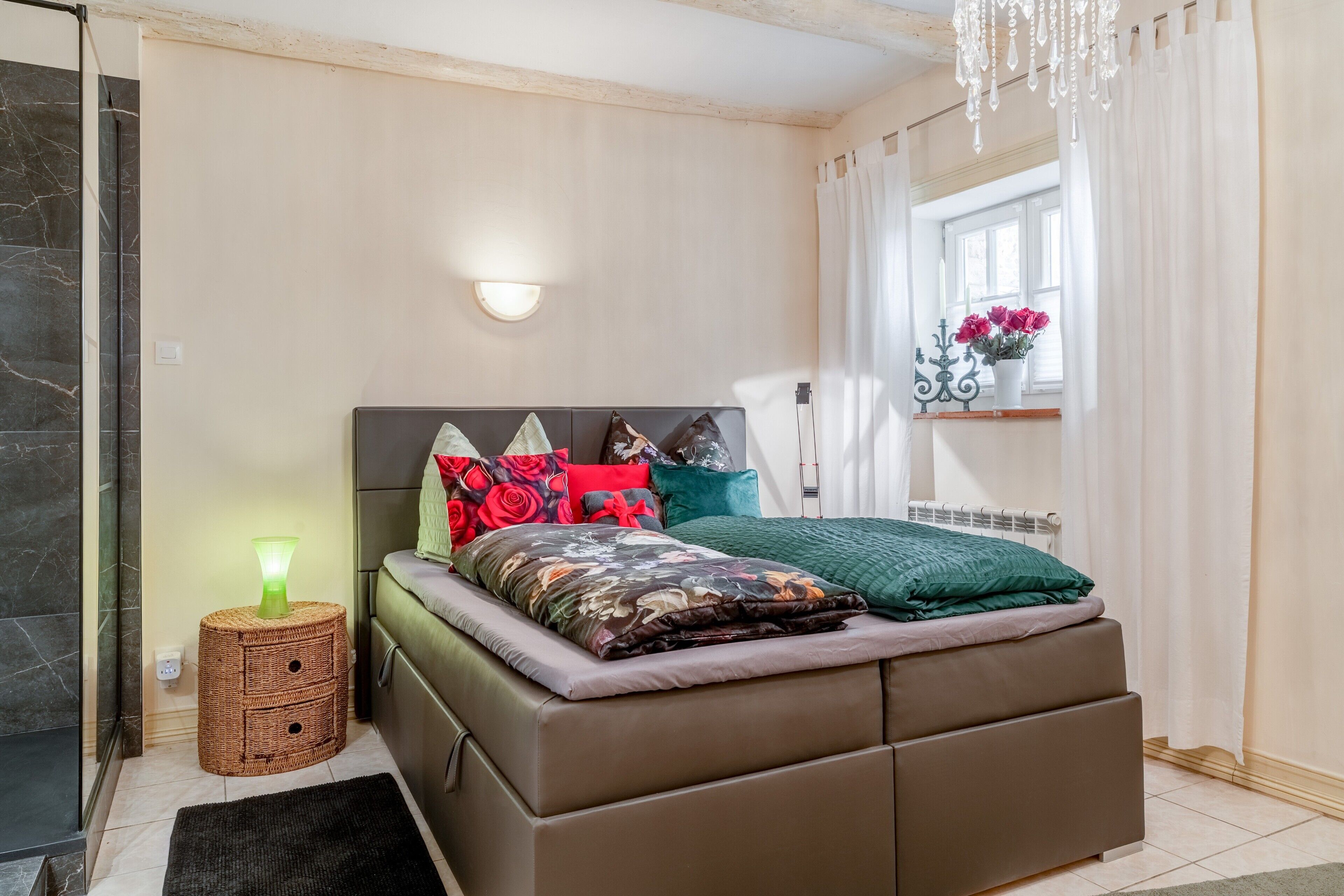 A bedroom with a bed, bedside table, wall-mounted light, window with curtains, and a small window with flowers.