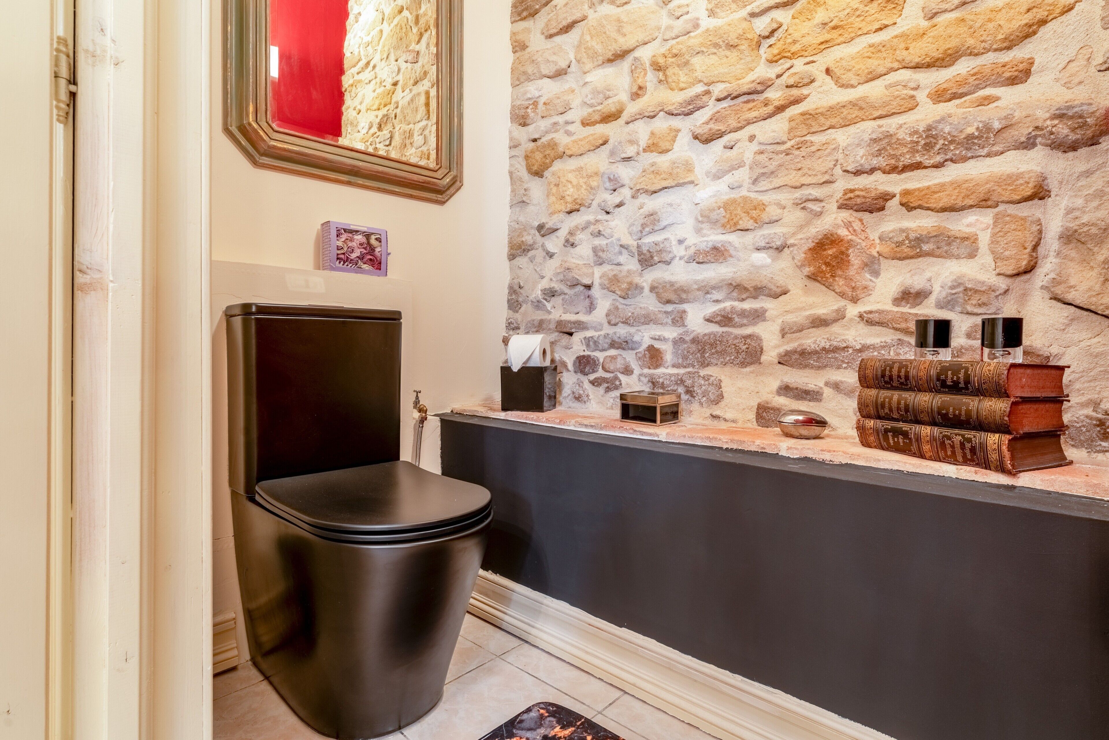 A bathroom with a stone wall, a black toilet, and a shelf with books and bottles.