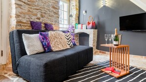 Un salon chaleureux avec un mur en pierre, un canapé agrémenté de coussins à motifs, une cheminée, un téléviseur et une petite table en bois sur laquelle se trouvent une bouteille et des verres.