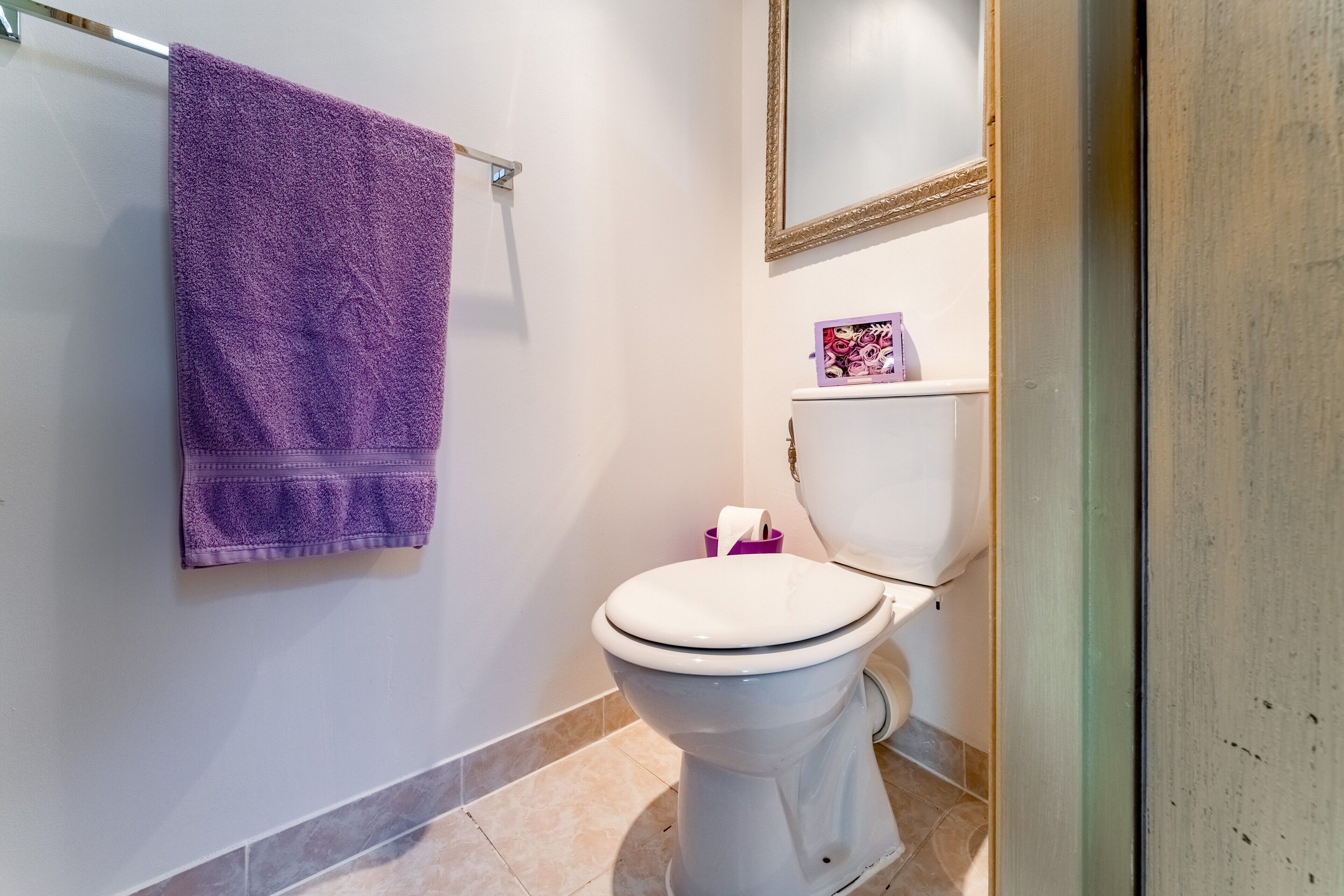 Ein Badezimmer mit einer weißen Toilette, einem violetten Handtuch an einem Halter, einer kleinen Ablage mit einem rosafarbenen Gegenstand und einem Spiegel über der Toilette.