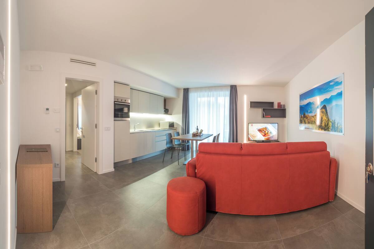 A modern living room with a red sofa and ottoman, a kitchen in the background, and a dining area.