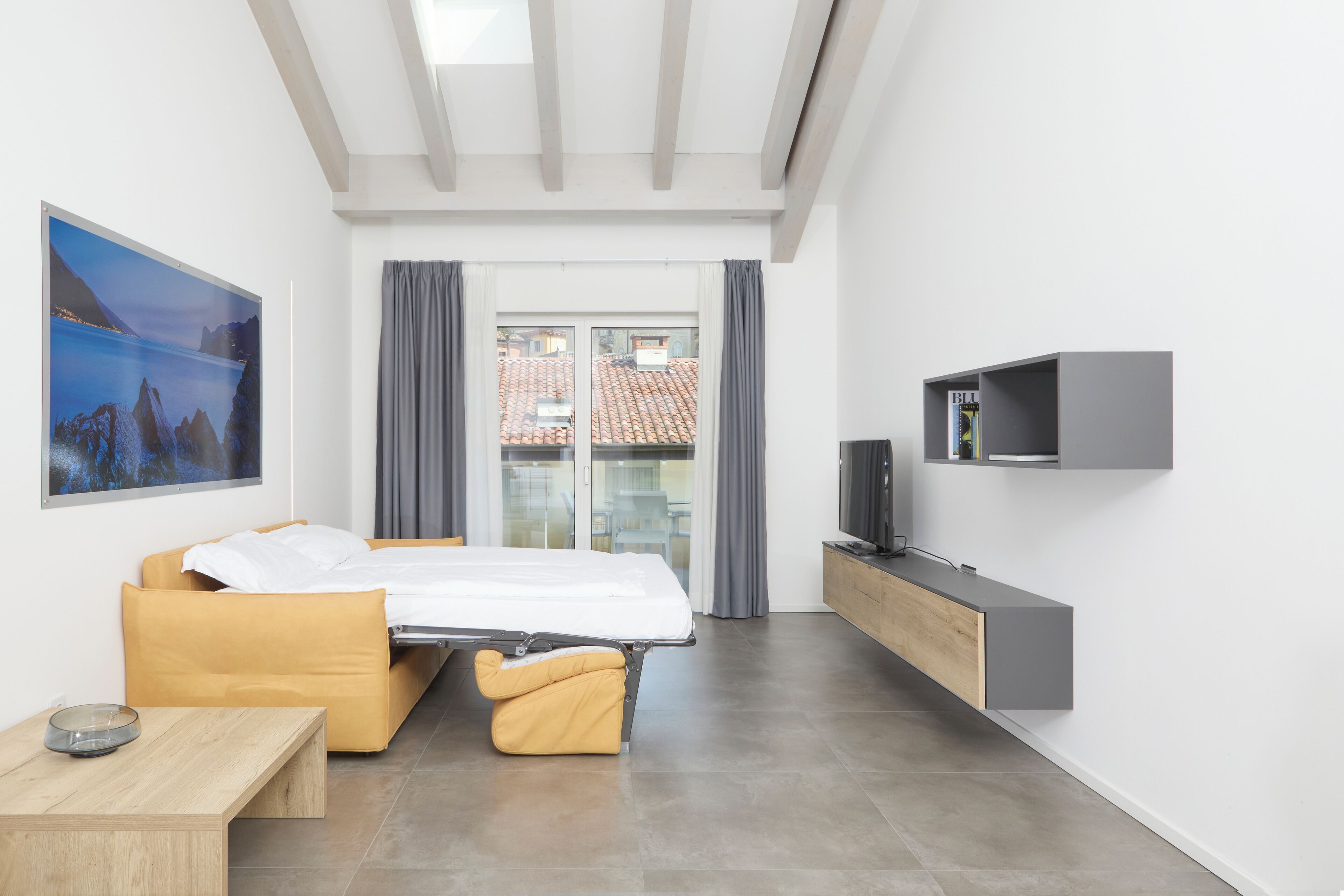 A modern hotel room with a bed, a small table, a TV, and a view of the outside.