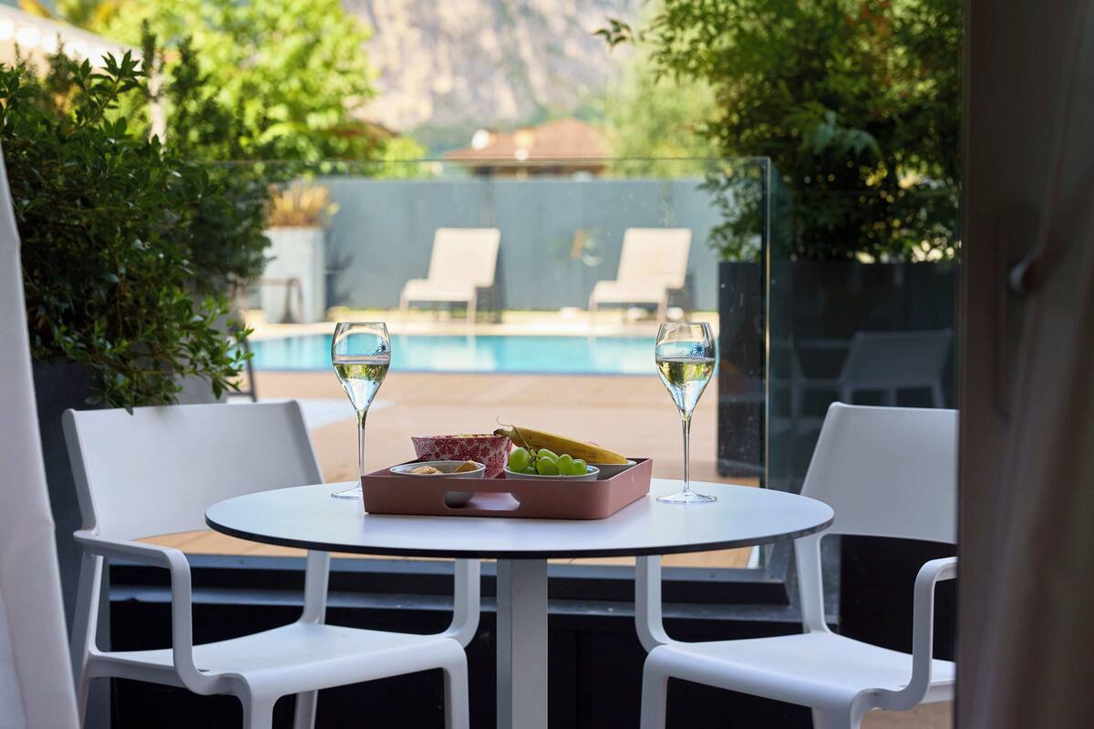 A table with a fruit tray and two glasses of white wine, overlooking a pool and lounge chairs.