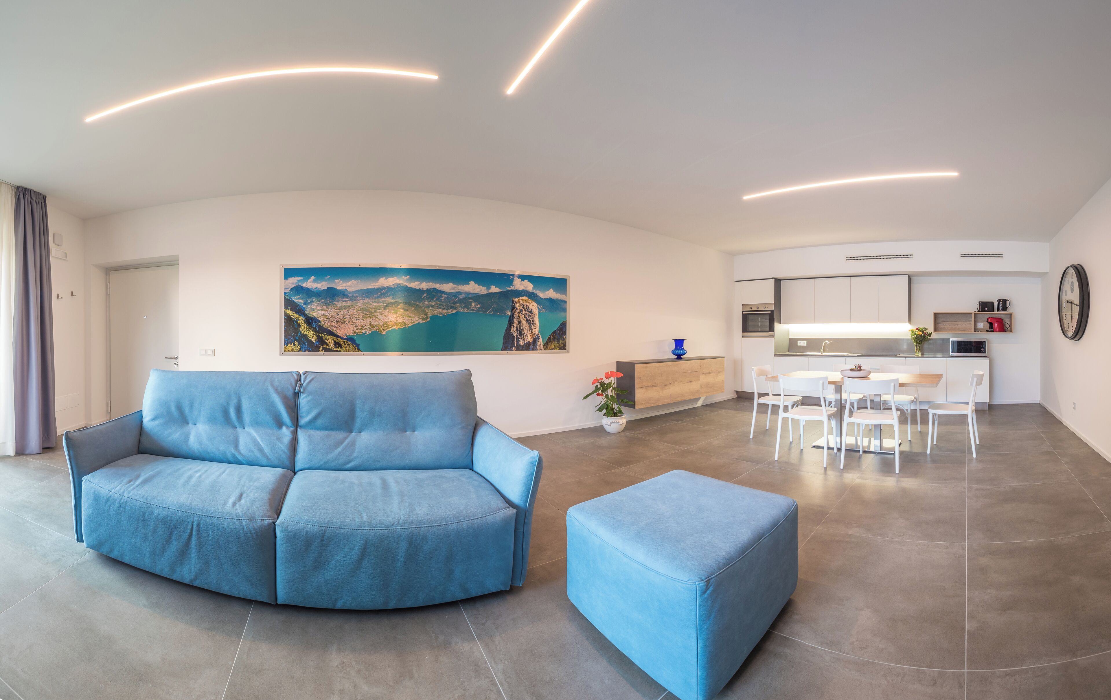 A modern living room with a blue sofa and ottoman, a dining area with white chairs, a kitchen with white cabinets, and a large landscape painting on the wall.