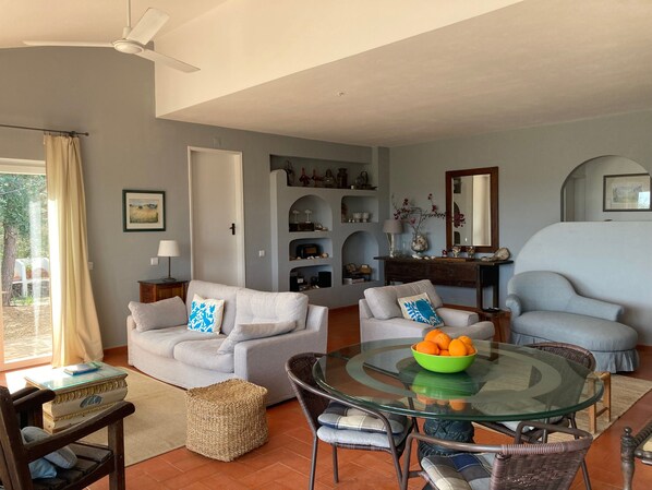 A living room with a glass table, two sofas, a wicker chair, and a dining table with a bowl of fruit.