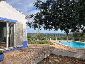 A poolside area with a view of the countryside, a white building with a blue trim, and a tree providing shade.